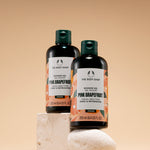 Two bottles of The Body Shop Pink Grapefruit Shower Gel 250 ML positioned on decorative stones against a soft beige backgroun