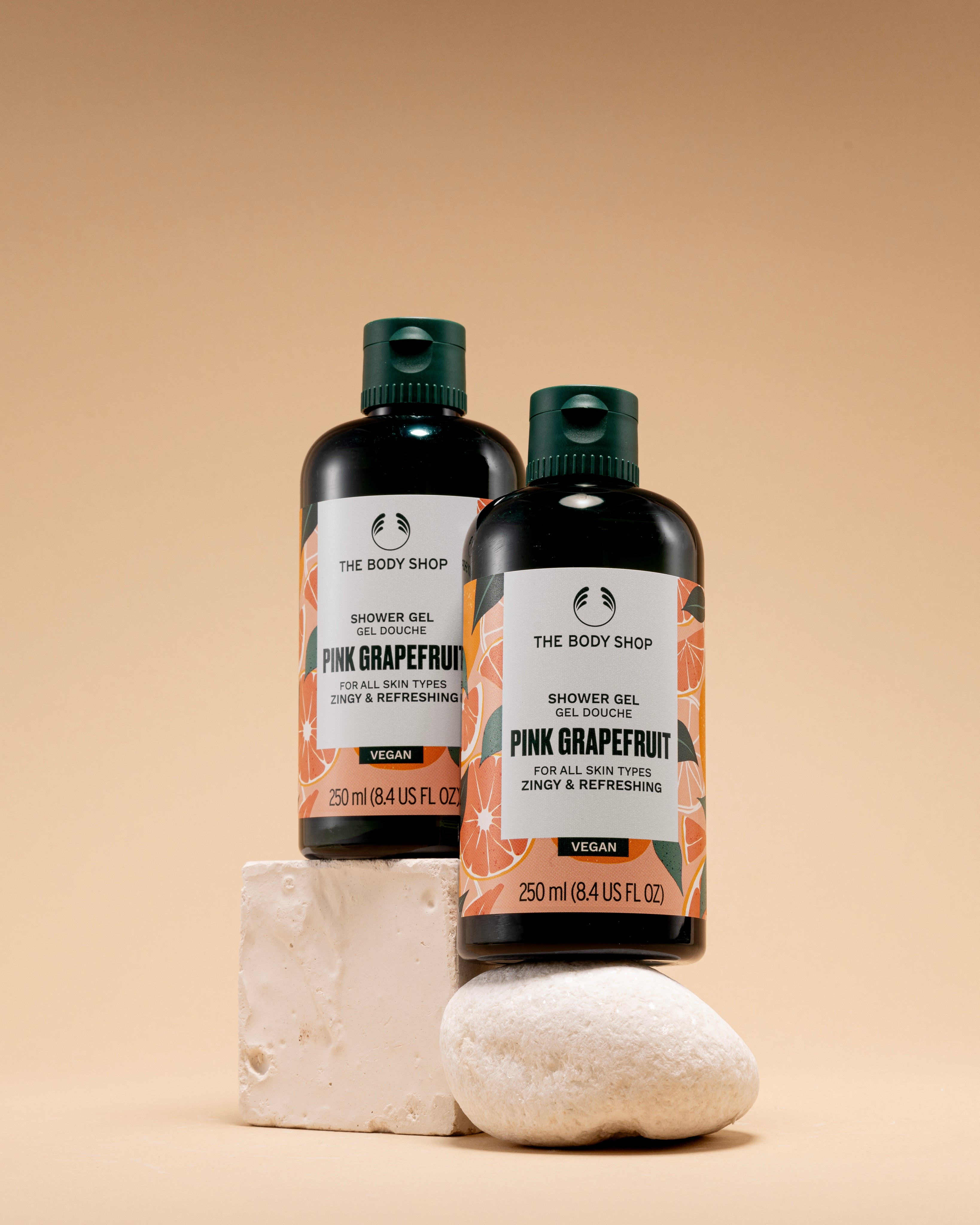 Two bottles of The Body Shop Pink Grapefruit Shower Gel 250 ML positioned on decorative stones against a soft beige backgroun