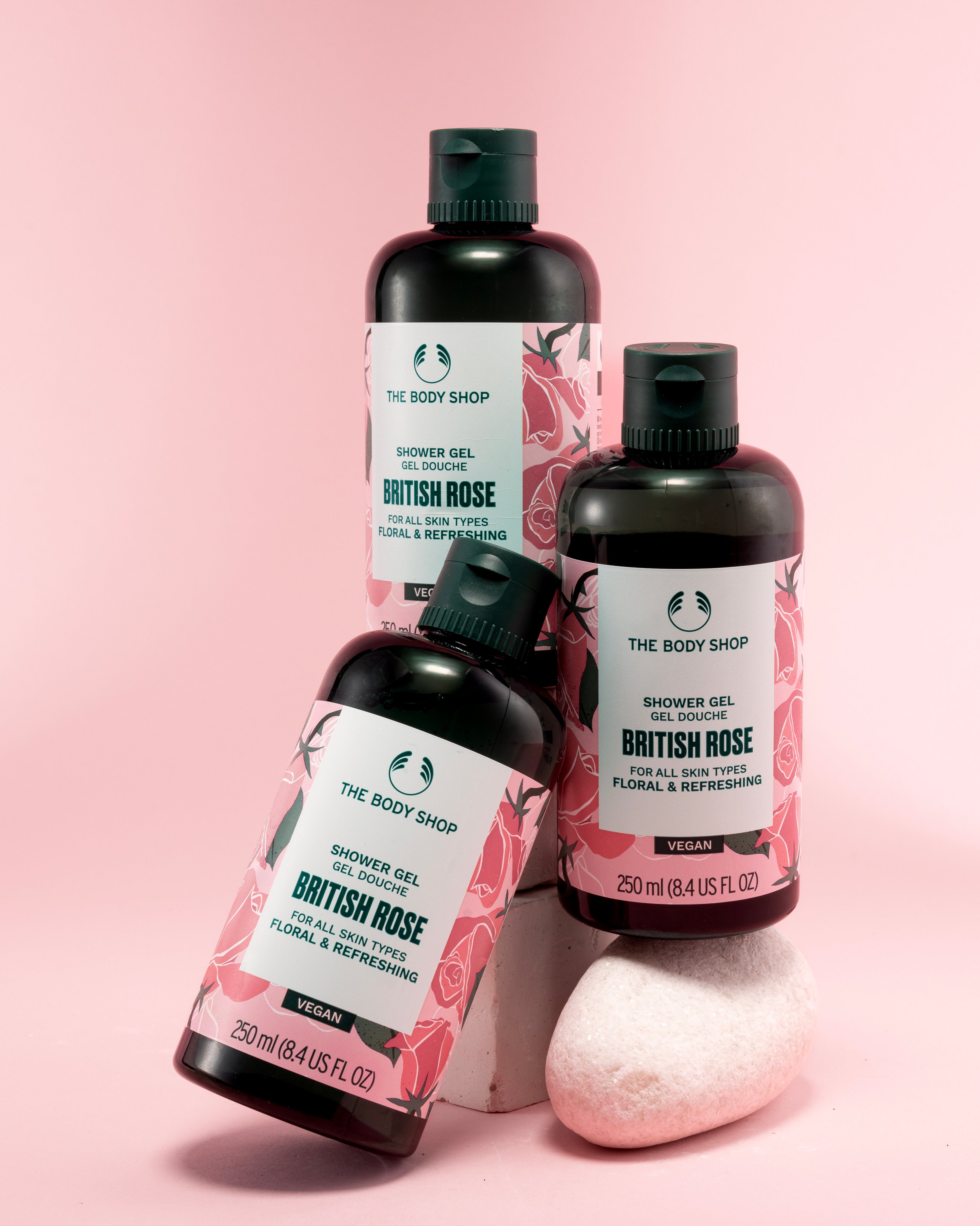 Three bottles of The Body Shop British Rose Shower Gel 250 ML stacked on a pink backdrop with decorative stone
