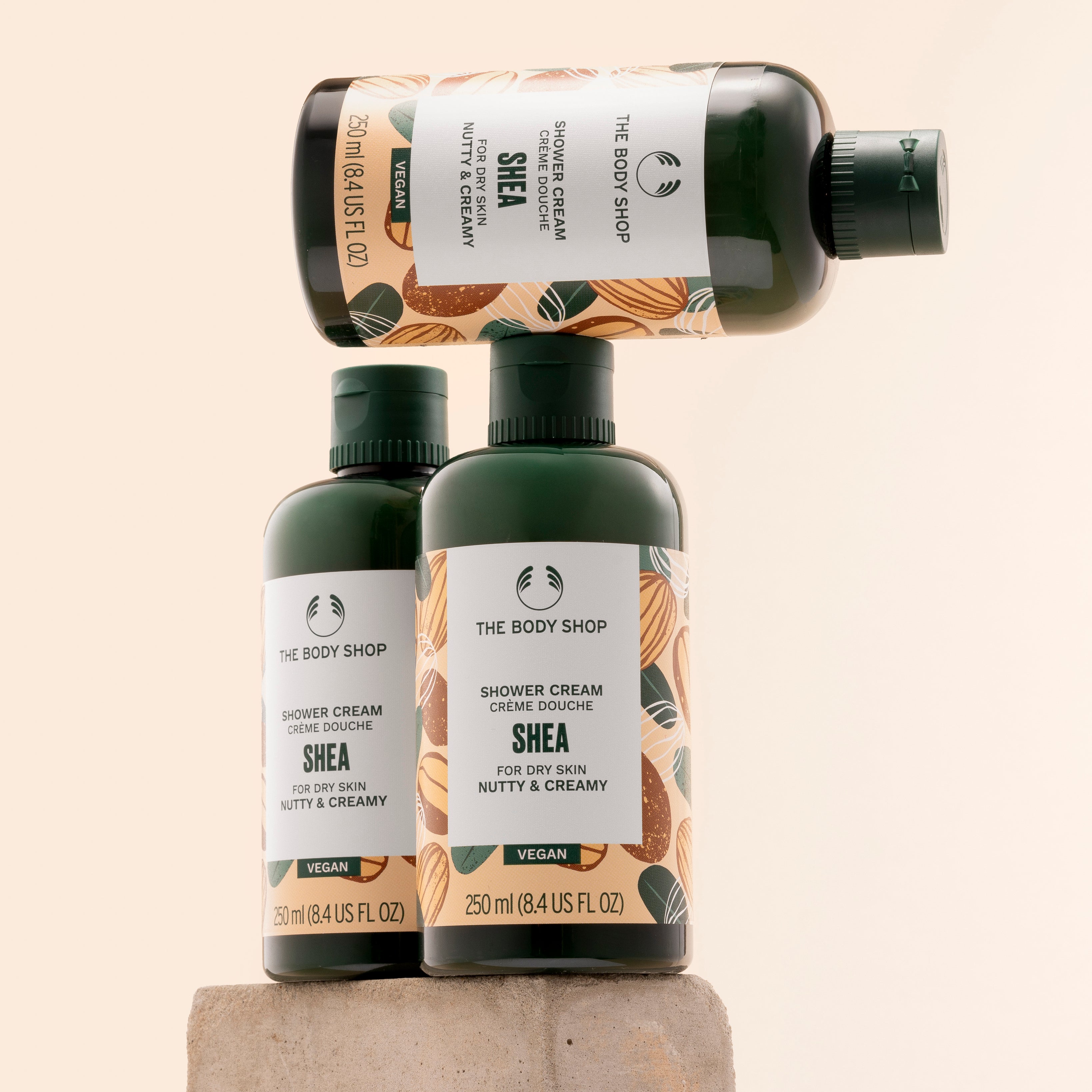 Three bottles of The Body Shop Shea Shower Cream 250ml elegantly stacked on a textured stone pedestal against a soft pastel b