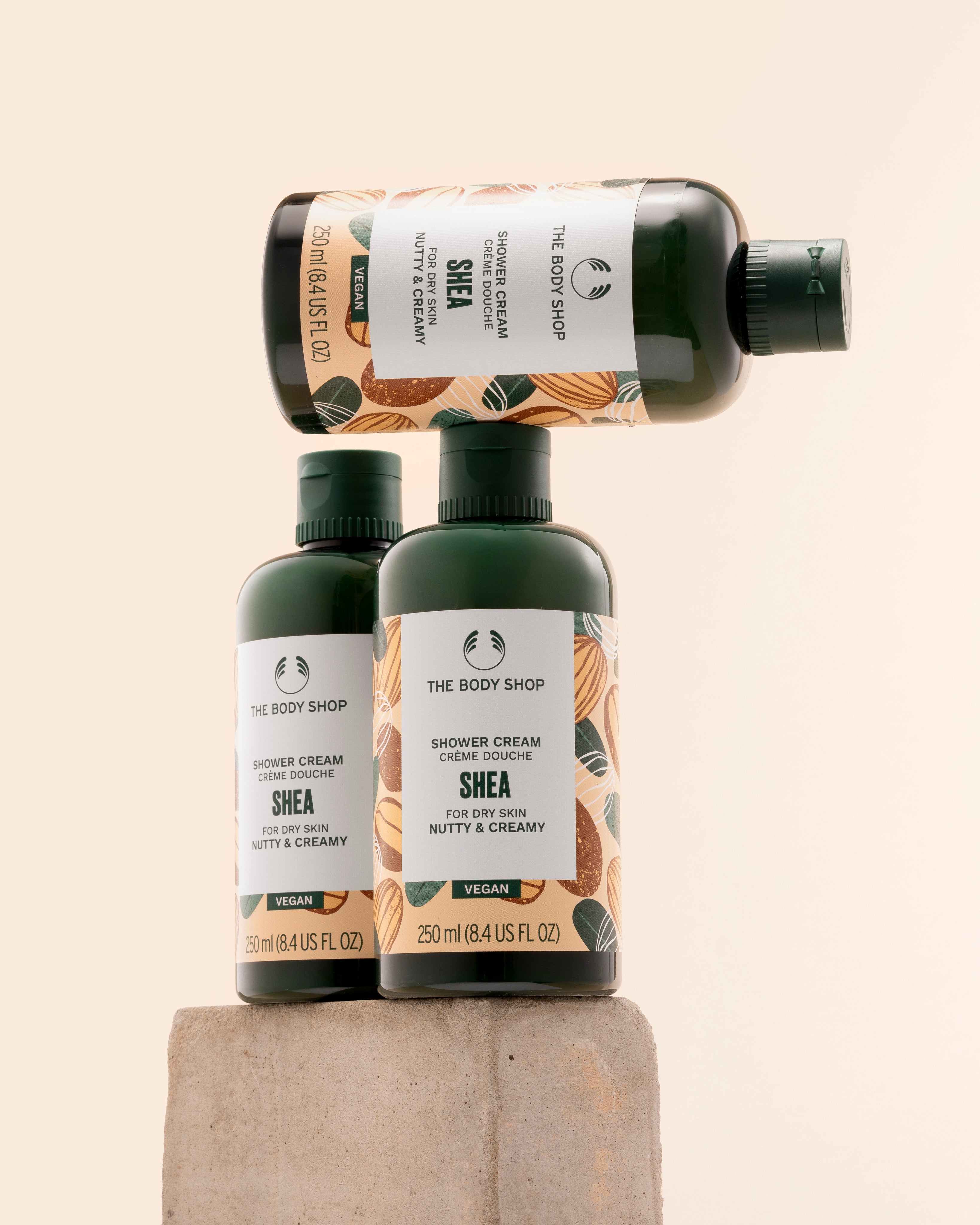 Three bottles of The Body Shop Shea Shower Cream 250ml elegantly stacked on a textured stone pedestal against a soft pastel b