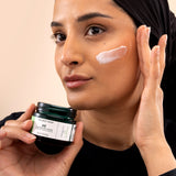 Woman applying The Body Shop Aloe Soothing Day Cream 50ML to her cheek while holding the jar close to her face.