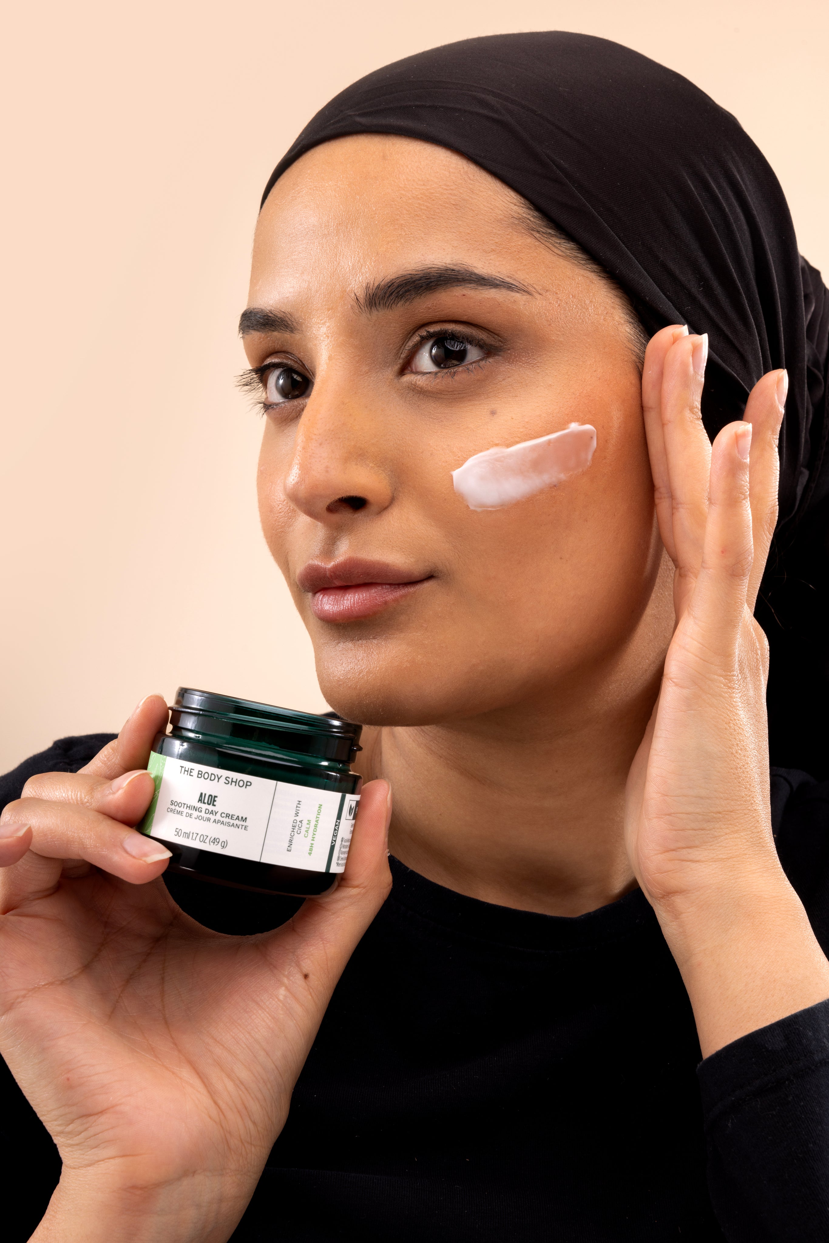 Woman applying The Body Shop Aloe Soothing Day Cream 50ML to her cheek while holding the jar close to her face.