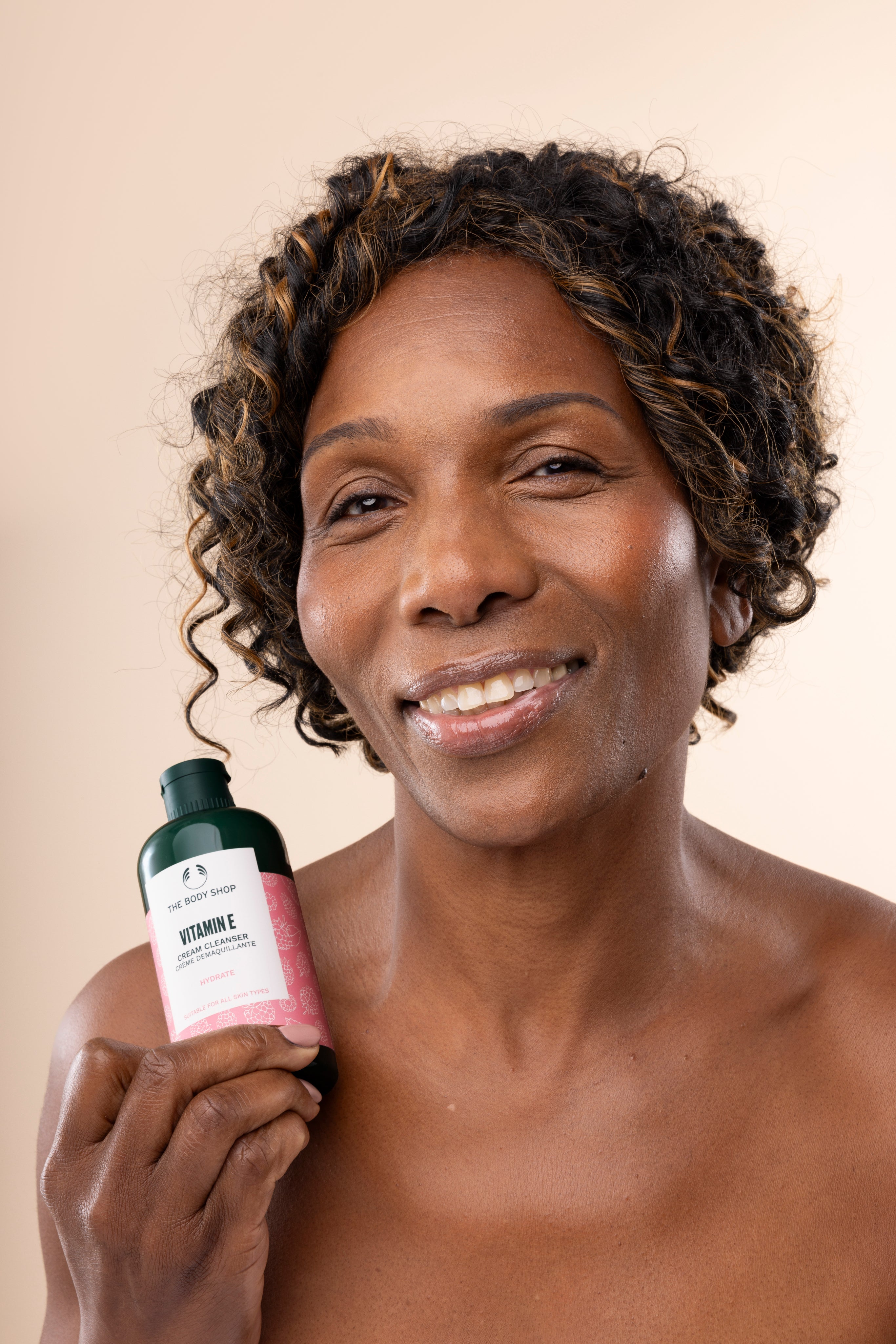 Woman smiling while holding The Body Shop Vitamin E Cream Cleanser 250 ML against soft, neutral background