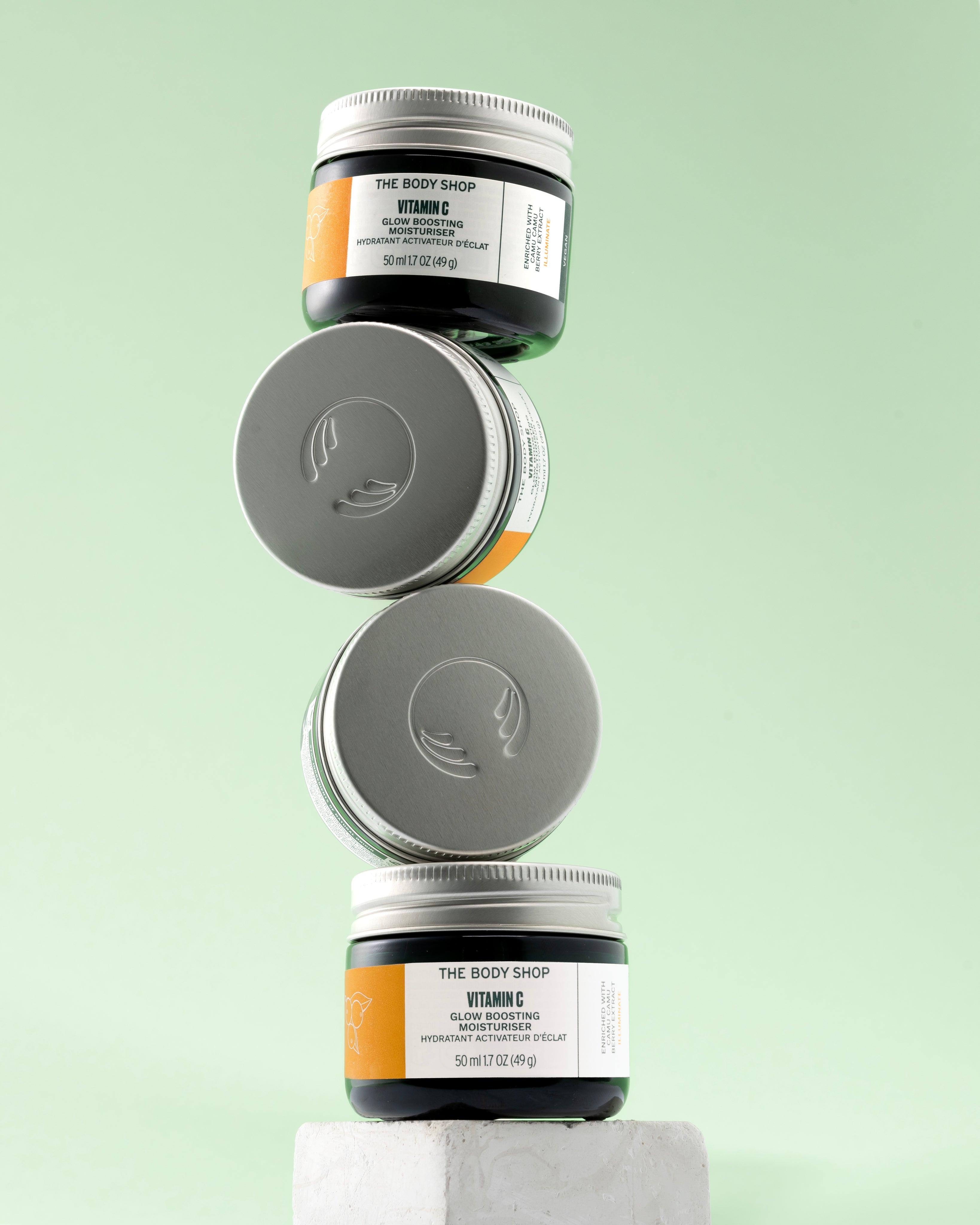 Three jars of The Body Shop Vitamin C Glow Boosting Moisturiser 50 ML stacked on a textured surface against a soft green back