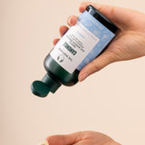 Hands pouring The Body Shop Camomile Gentle Eye Makeup Remover 250 ML onto a cotton pad against a soft, neutral background