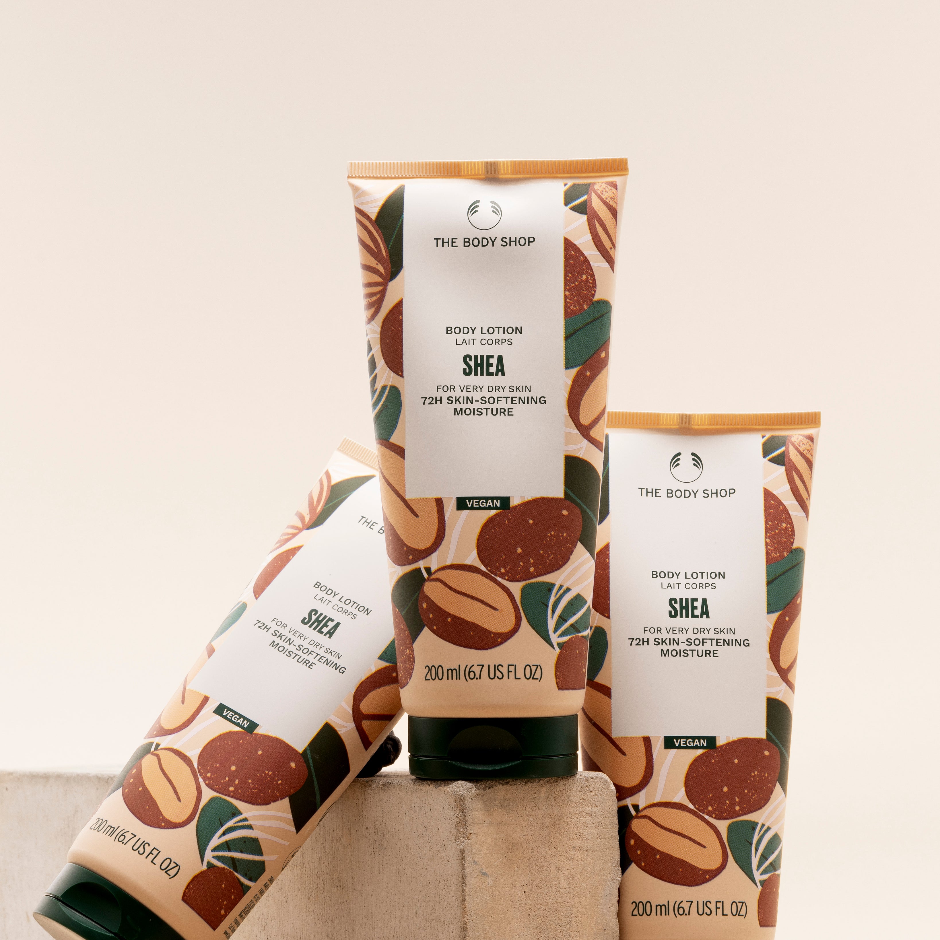 Three tubes of The Body Shop Shea Body Lotion 200 ML artistically arranged on a stone surface against a soft, muted backgroun