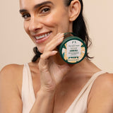 Woman holding The Body Shop Almond Milk Body Yogurt 200ml in front of her face against a studio backdrop