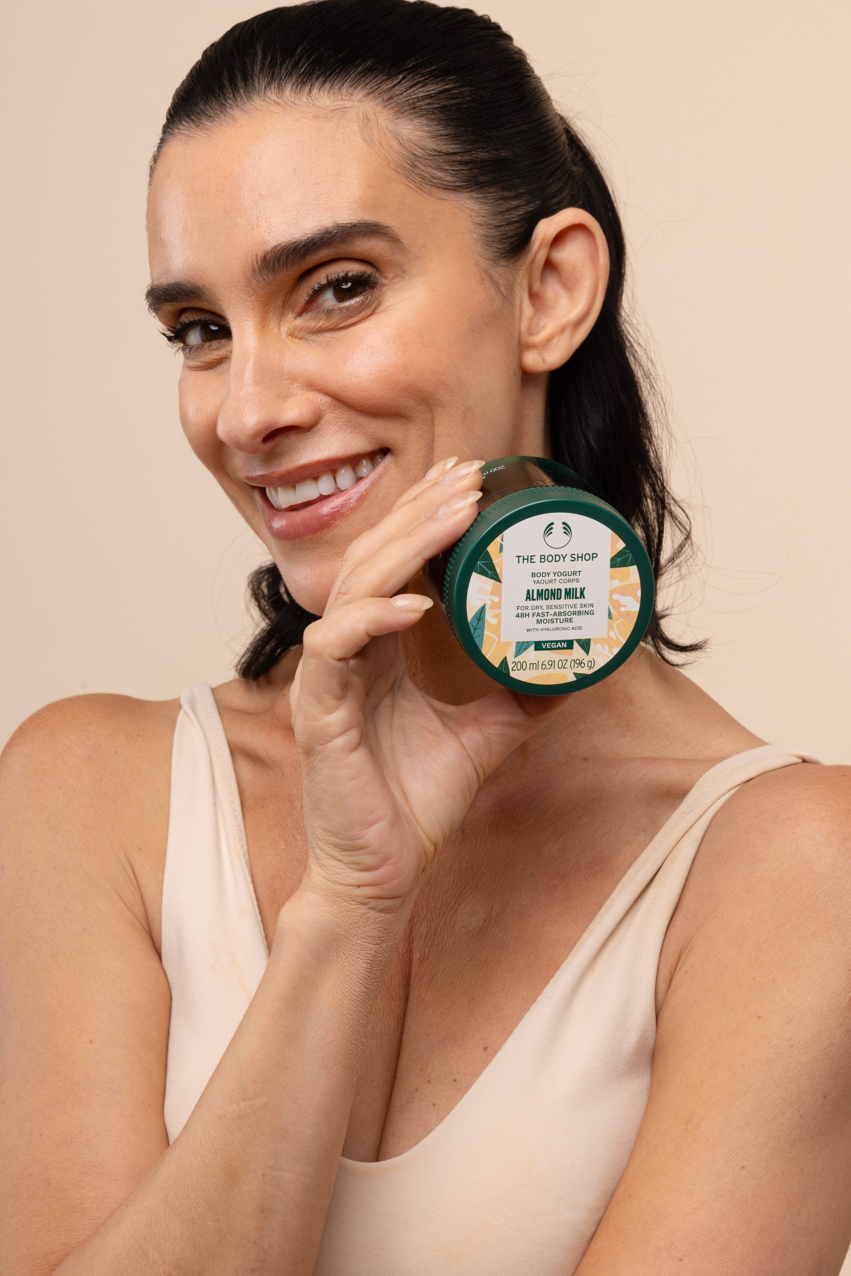 Woman holding The Body Shop Almond Milk Body Yogurt 200ml in front of her face against a studio backdrop