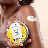 Woman applying The Body Shop Mango Body Butter 200 ML on her arm while holding the container in a bright setting.