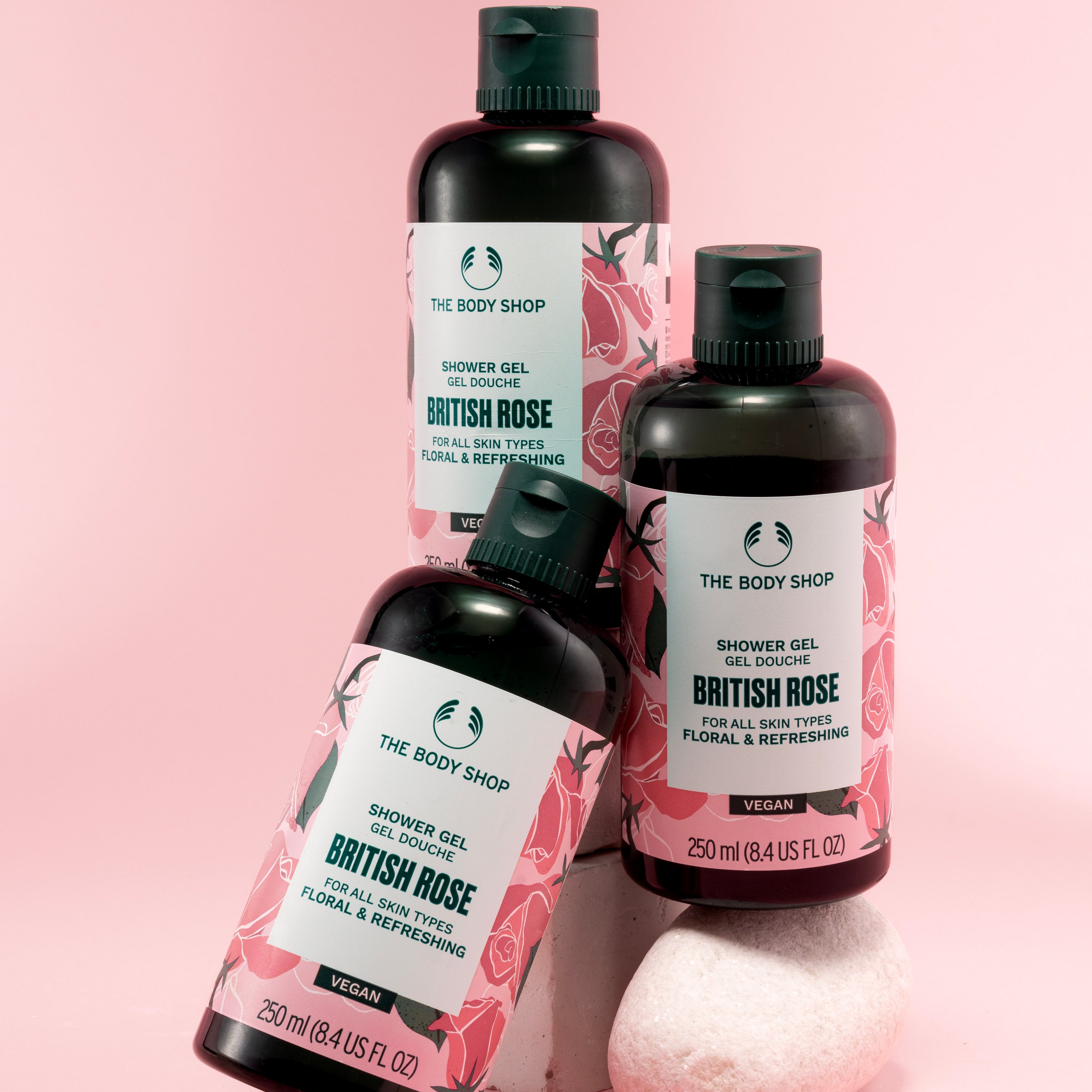 Three bottles of The Body Shop British Rose Shower Gel 250 ML stacked on a pink backdrop with decorative stone