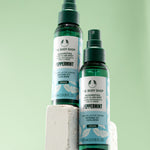 Two bottles of The Body Shop Peppermint Invigorating Foot & Leg Mist 100 ML on stone pedestal against soft green background