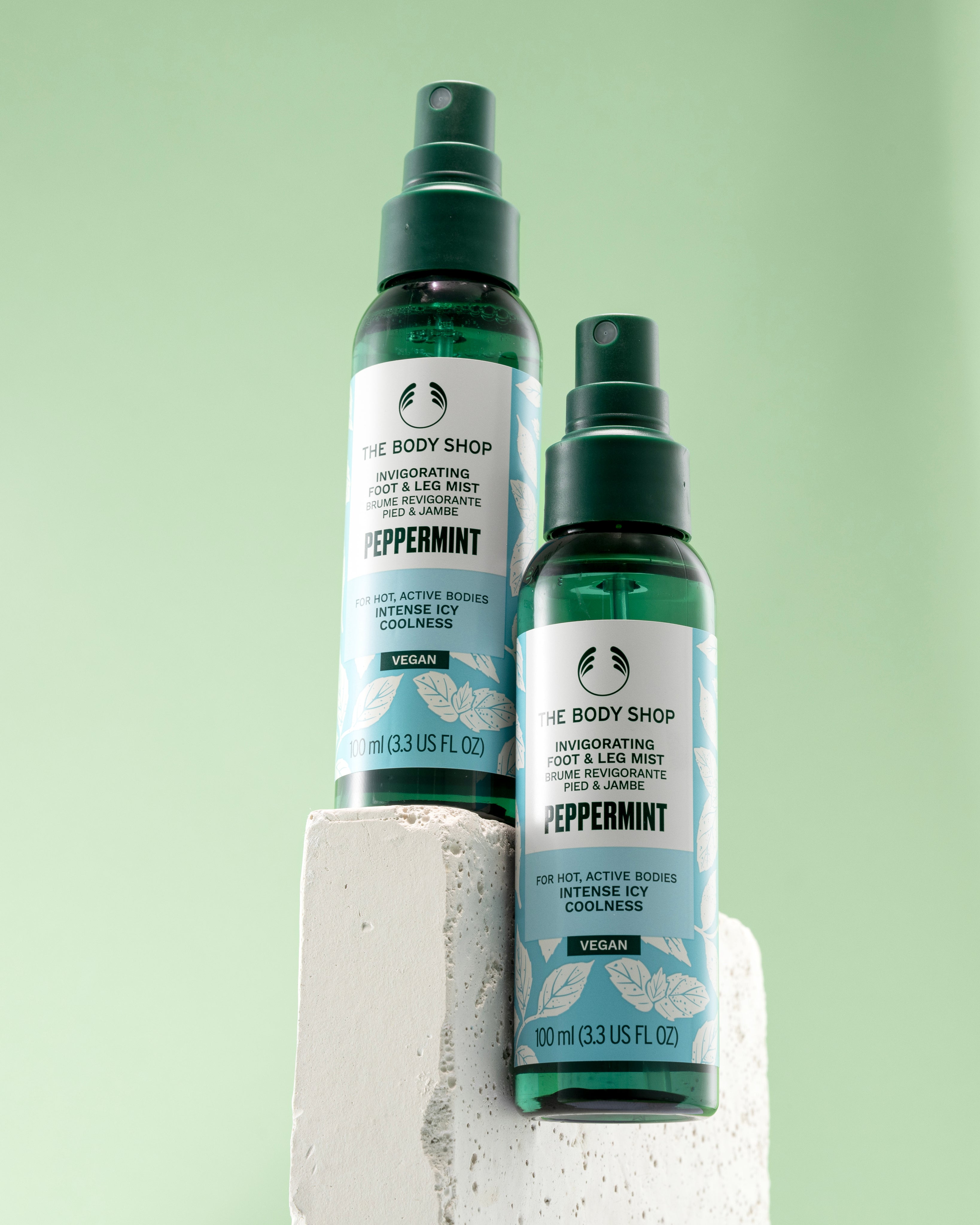 Two bottles of The Body Shop Peppermint Invigorating Foot & Leg Mist 100 ML on stone pedestal against soft green background