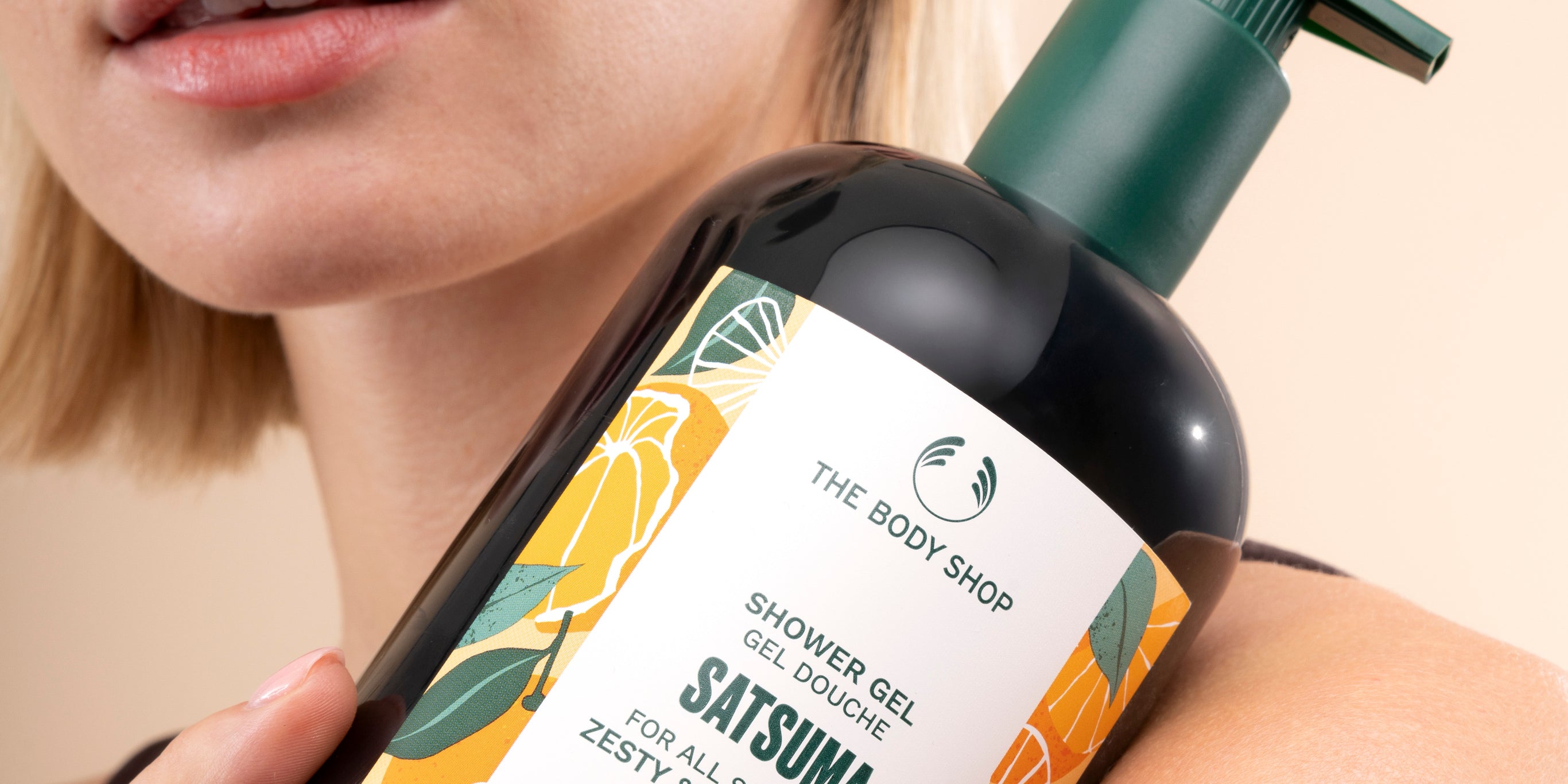 Young woman holding The Body Shop Satsuma Shower Gel 750 ML showcasing the product against her arm in a natural setting.