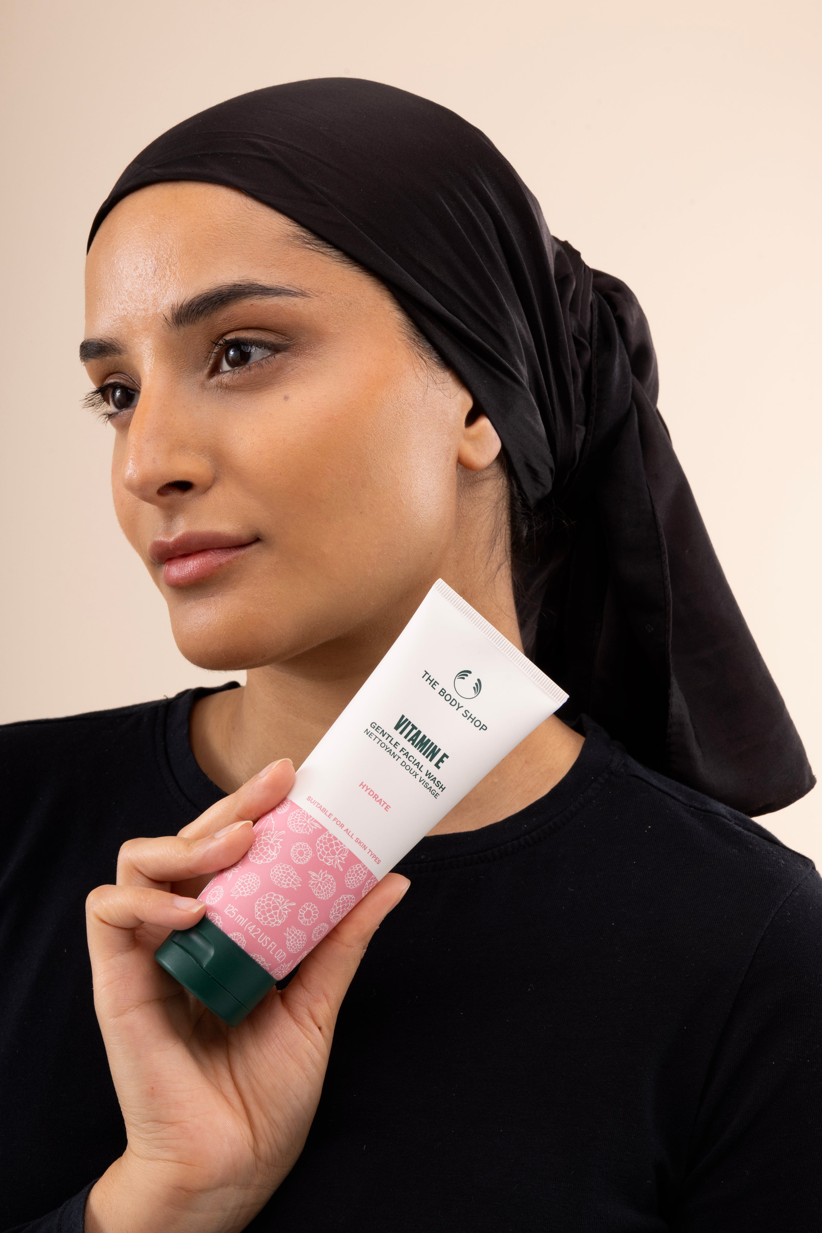 Woman holding The Body Shop Vitamin E Gentle Facial Wash 125ml against soft pastel background highlighting hydration.