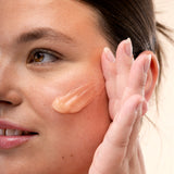 A person applies a cream to their cheek against a plain background.