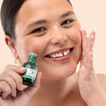 Woman applying The Body Shop Tea Tree Oil 20 ML, with green bottle, in bathroom setting