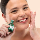 Woman applying The Body Shop Tea Tree Oil 20 ML, with green bottle, in bathroom setting