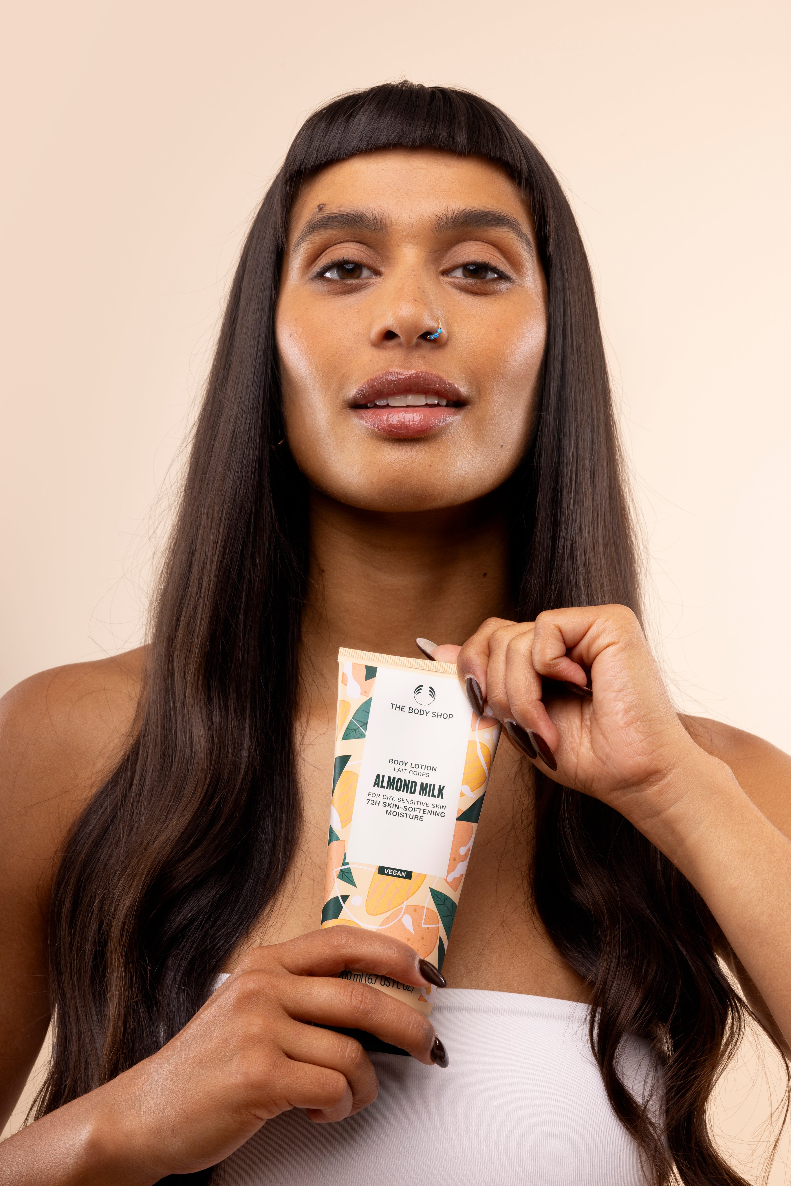 Woman holds The Body Shop Almond Milk Body Lotion 200ml against soft peach-toned background