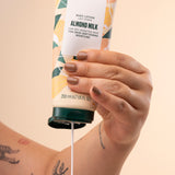 Hands dispensing The Body Shop Almond Milk Body Lotion 200 ML, showcasing creamy texture on white studio backdrop