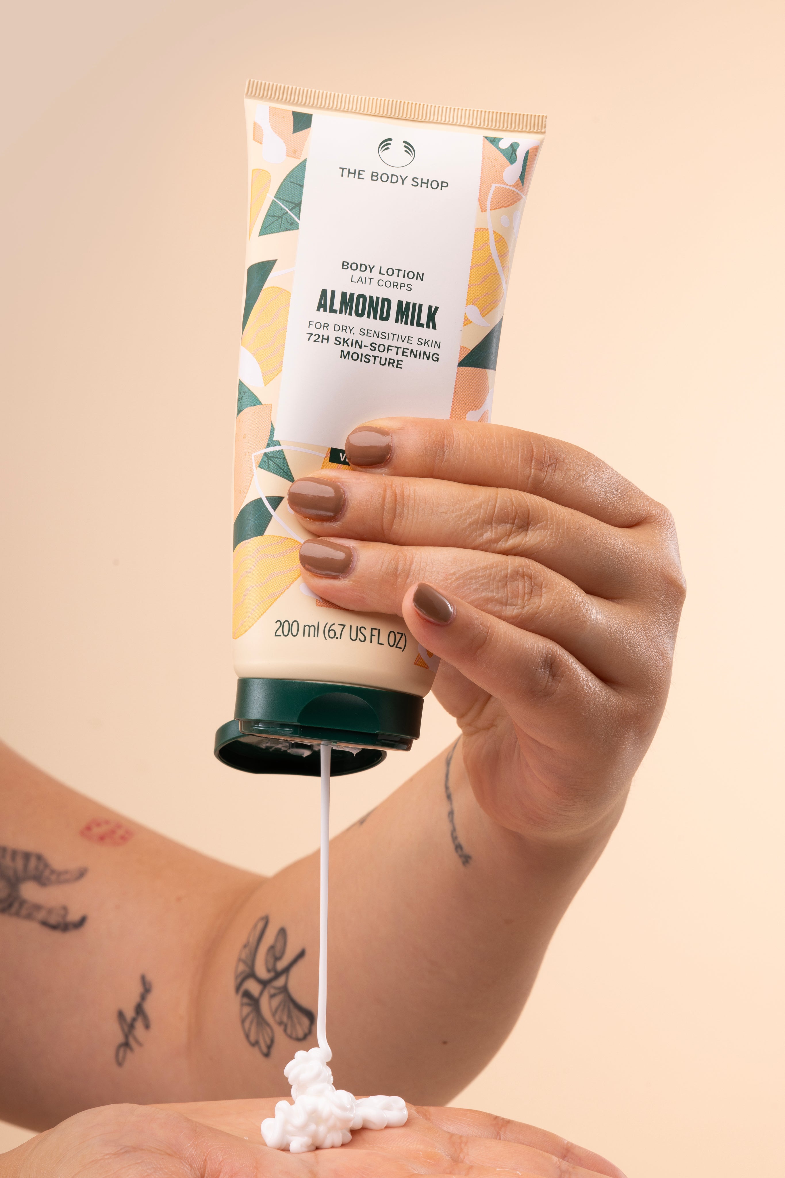 Hands dispensing The Body Shop Almond Milk Body Lotion 200 ML, showcasing creamy texture on white studio backdrop