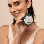 Woman holding The Body Shop Almond Milk Body Yogurt 200ml in front of her face against a studio backdrop