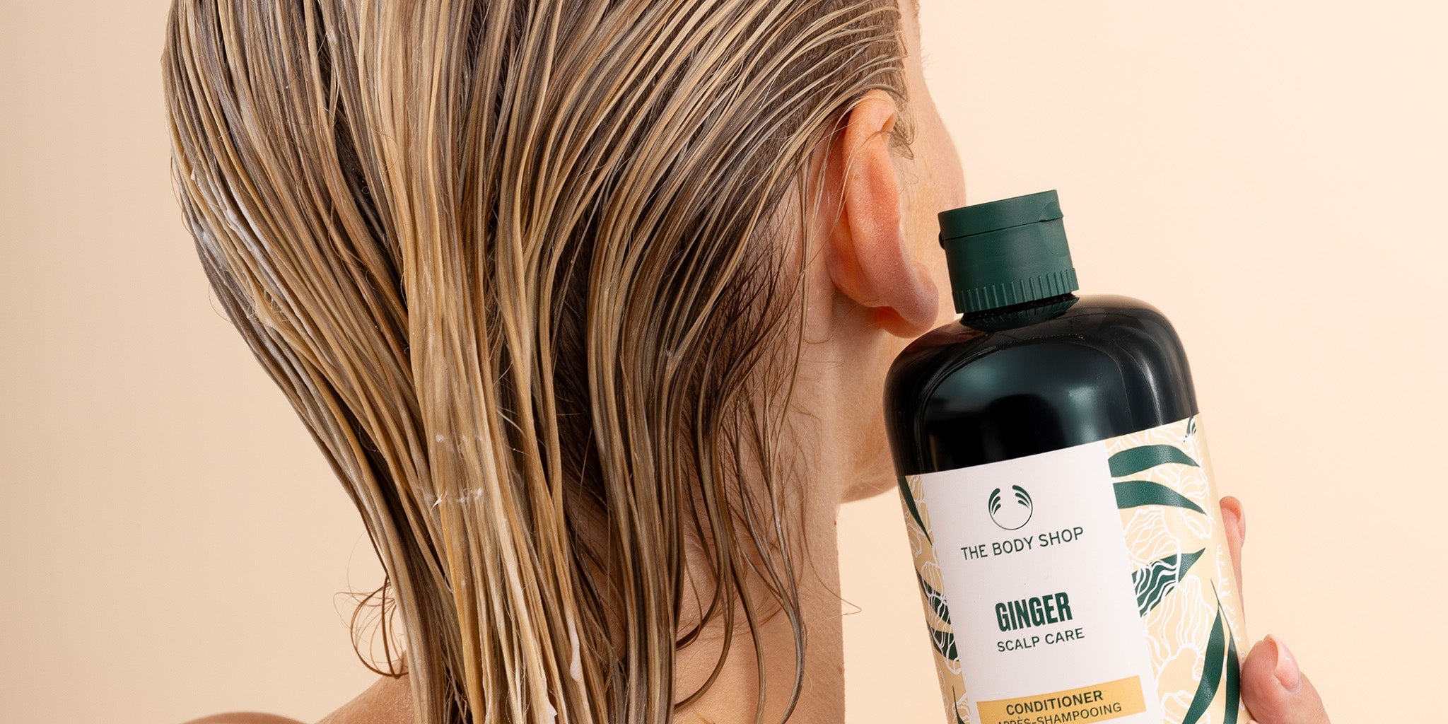 Person with wet hair holding a bottle of The Body Shop Ginger Conditioner against a beige background