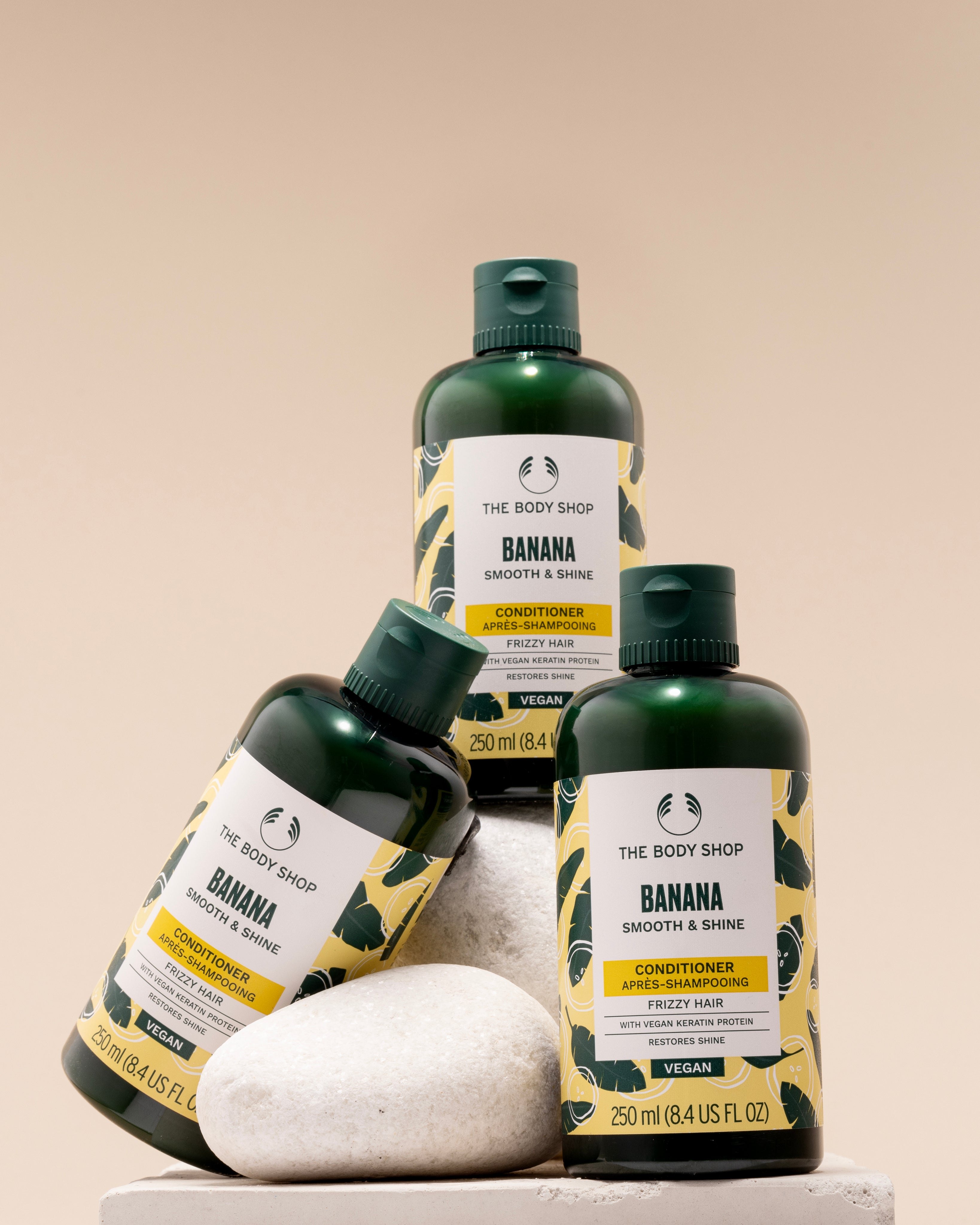 Three bottles of The Body Shop Banana Smooth & Shine Conditioner 250 ML displayed on a pedestal of stones against soft beige