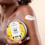 Woman applying The Body Shop Mango Body Butter 200 ML on her arm while holding the container in a bright setting.