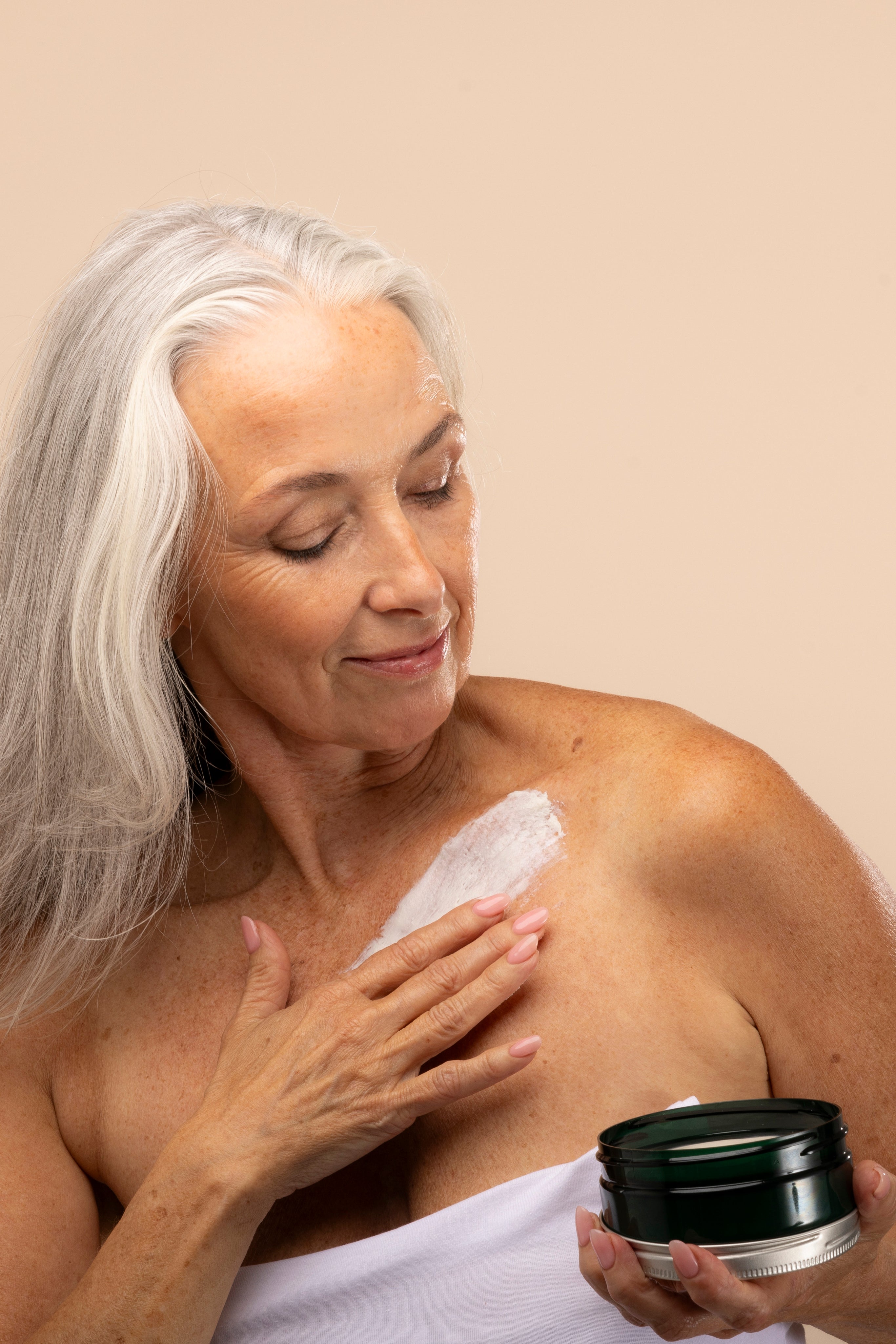 Elderly woman applying Shea Body Butter 200ml against studio backdrop, showcasing its creamy texture on her shoulder.